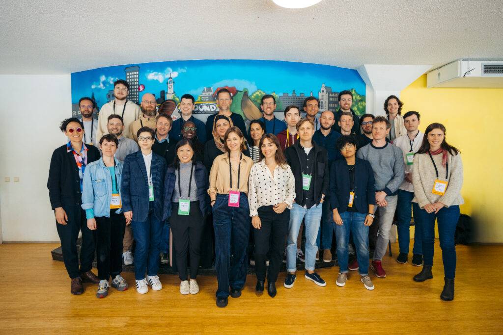 A group photo of 27 people standing together in a room with a colorful cityscape mural on the wall behind them.