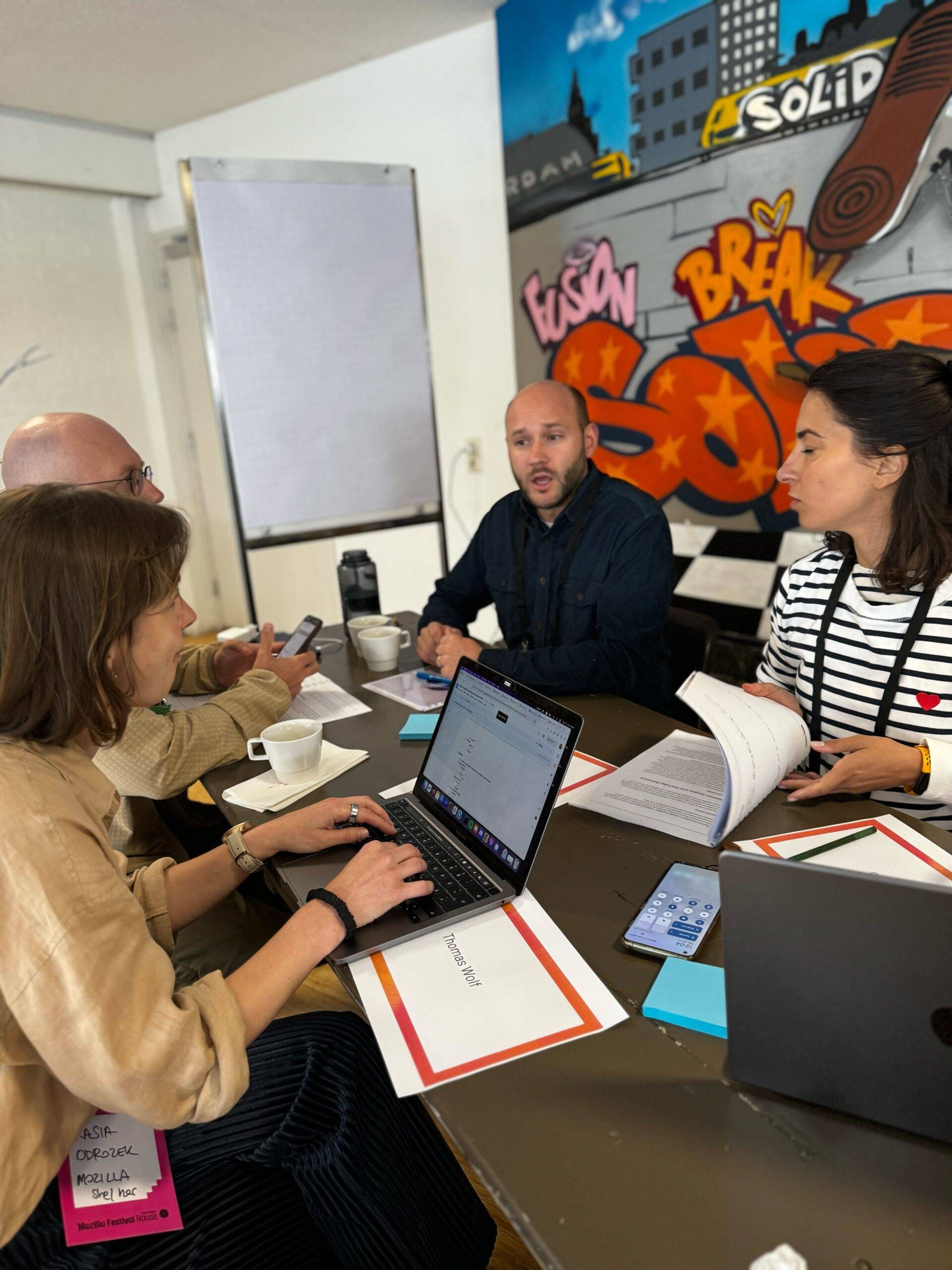 A group of four people sitting around a table with laptops and documents, engaged in a discussion. One person types on a laptop, while others look at papers and a phone. A colorful graffiti mural is on the wall behind them.